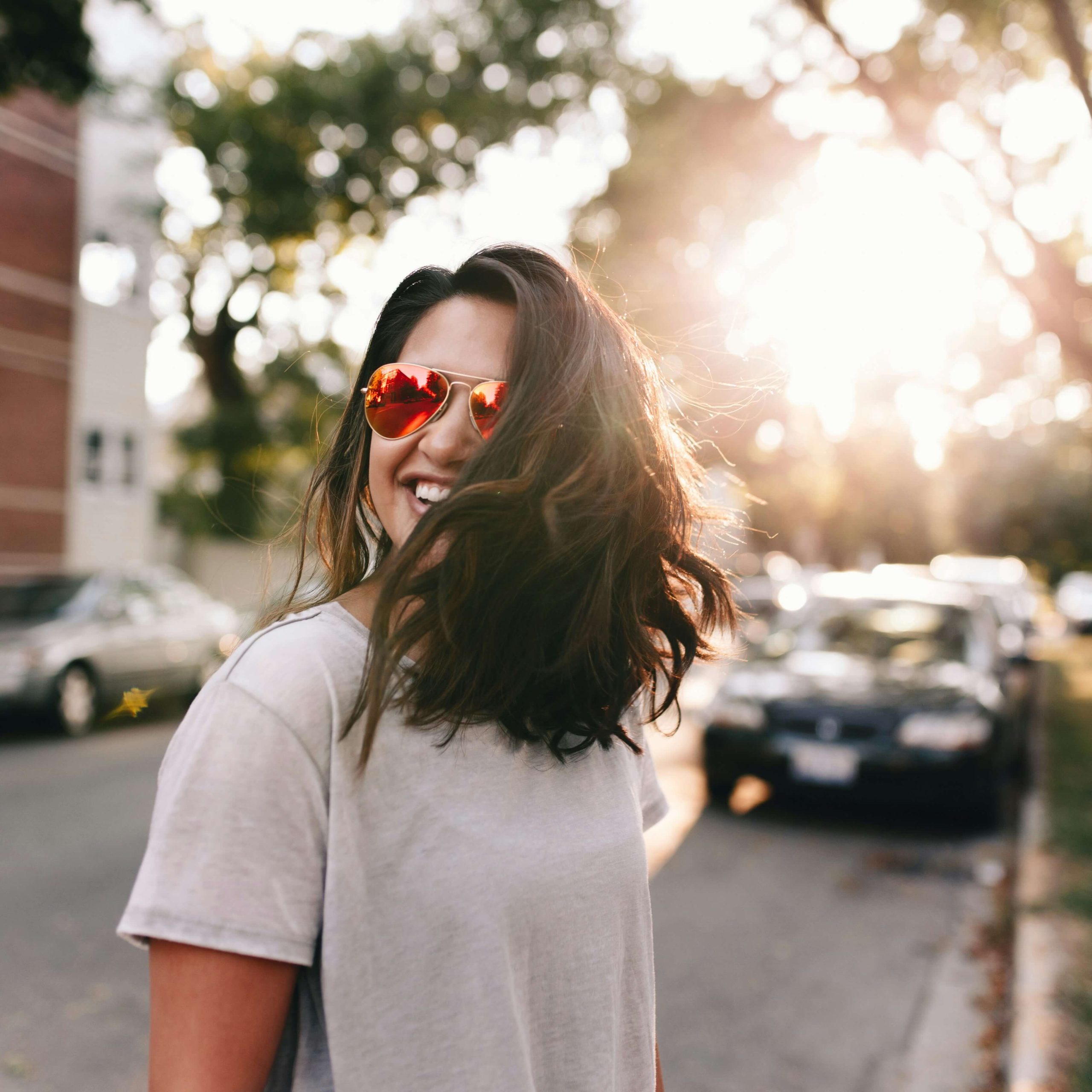 Portrait d'une jeune femme en extérieur. Brune, les cheveux mi-long, portant des lunettes de soleil, et rayonnant.
