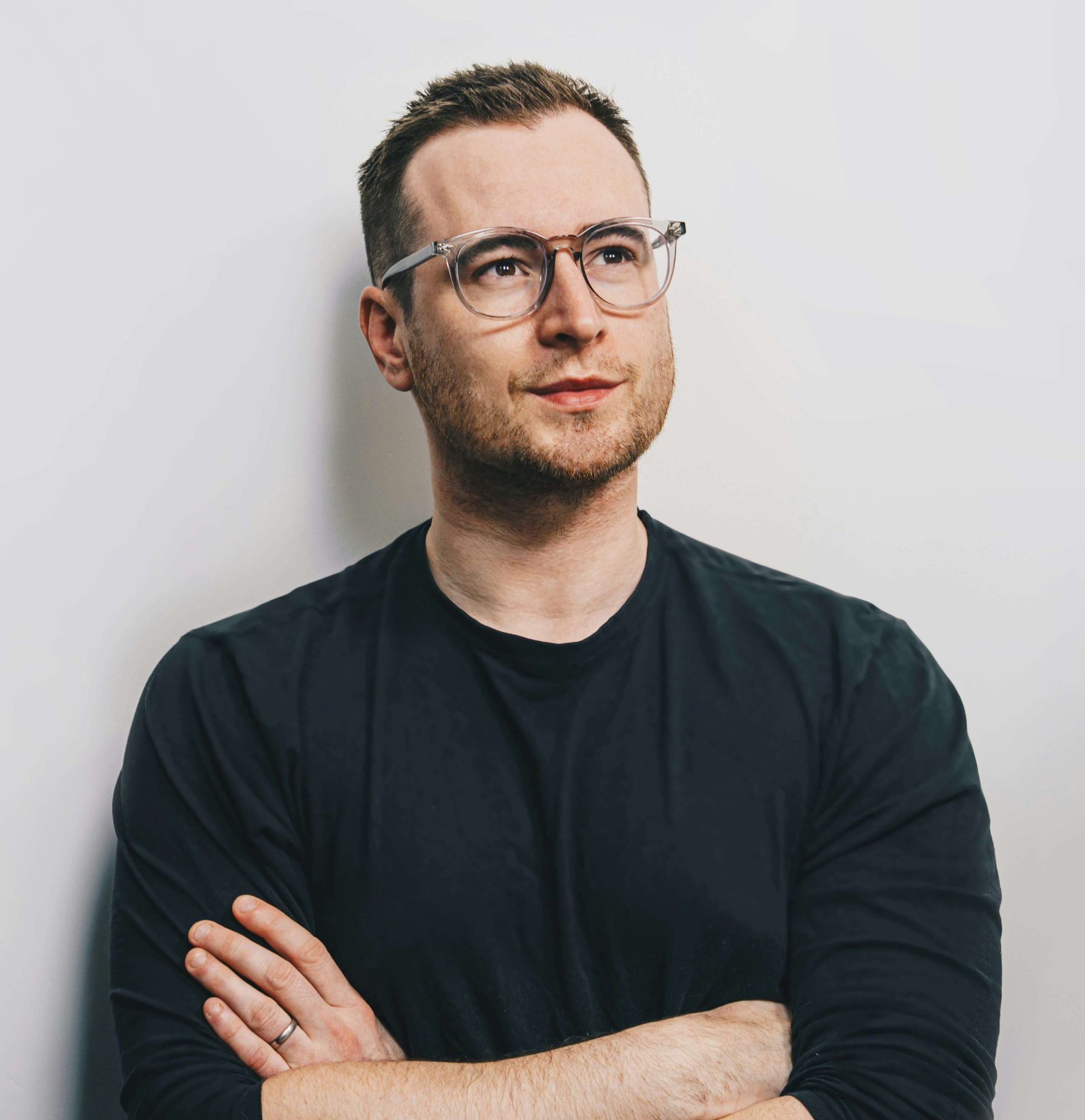Un homme brun au lunette, portant un t-shirt noir mange longue, les bras croisés. Un alliance à sa mains gauche, le regarde optimiste au loin.
