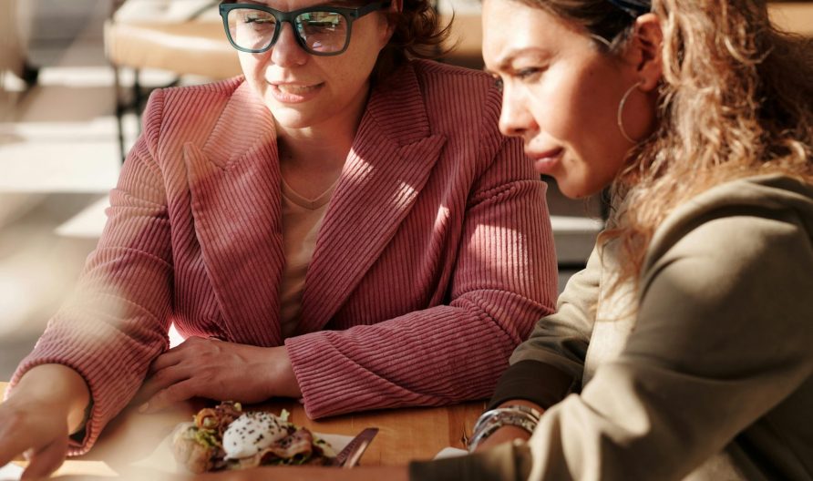 Photo de deux jeunes femmes en train de collaborer, les yeux river sur l'ordinateur. Elles sont dans un café, très lumineux.